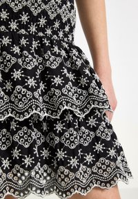 Abito in tessuto nero con ricami floreali bianchi, volants a strati e orlo scallop. Il design comprende motivi e texture intricate.