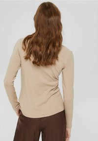 Camicia beige a maniche lunghe realizzata in tessuto morbido, con orlo arrotondato e silhouette aderente, indossata con pantaloni marroni.