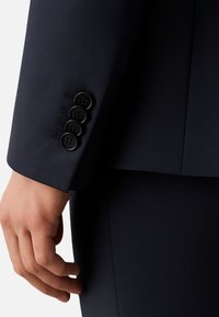 Manica di giacca blu navy con quattro bottoni neri, texture liscia, vestibilità su misura e linee pulite. Mano visibile, posizione rilassata.