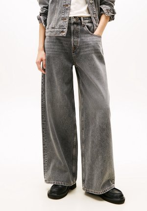 Personne portant un jean gris délavé à jambes larges, les mains dans les poches, associé à des chaussures noires et une veste en jean courte.