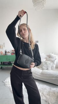 Femme en tenue noire tenant un sac bandoulière noir avec un logo, debout dans un salon lumineux avec un meuble vert et un canapé blanc.