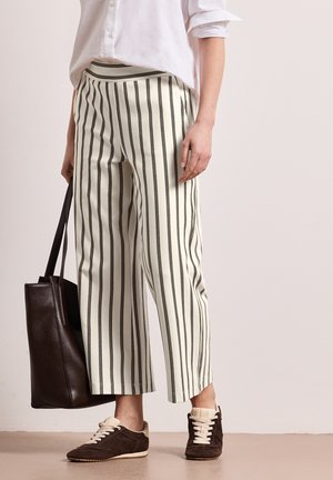 Osoba ubrana w białe spodnie w czarne pionowe paski, białą koszulę z podwiniętymi rękawami, ciemnobrązowe trampki, trzymająca ciemnobrązową torbę typu tote.