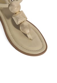 Beige sandaal met decoratieve gouden accenten, met drie textuurelementen: een kleine bol, een vierkant en een druppelvorm op de band.