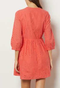 Robe corail avec des détails brodés, manches semi-transparentes, taille cintrée et ourlet évasé. Tissu en coton texturé et léger.
