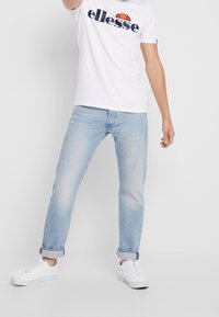 Bílé tričko s logem "ellesse", světle modré džíny a bílé tenisky. Džíny mají design s ohrnutým lemem, látka má vybledlou strukturu.