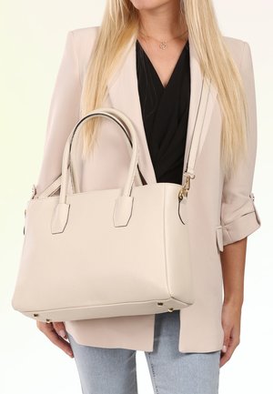 Frau in hellem Blazer und schwarzem Top hält große cremefarbene Lederhandtasche mit doppelten Griffen und Schulterriemen.