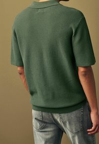 Mørkegrøn struktureret polo-shirt med kort krave, som har et tætsiddende kant og korte ærmer. Båret med lysegrå denim jeans.