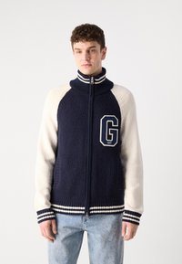 Zip-up pletený sveter s námorníckou telou, smotanovými rukávmi, vysokým golierom a rebrovanými manžetami so prúžkami. Predná nášivka s logom a textúrovaná látka.