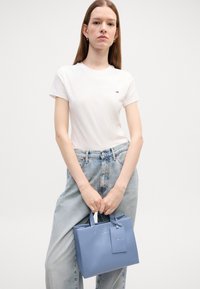 Bolso rectangular de color azul claro con asas cortas, textura brillante y una etiqueta. Mujer vestida con una camiseta blanca y jeans de denim claros.