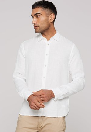 Mann mit kurzen dunklen Haaren, der ein weißes, langärmliges Hemd mit Knöpfen und beigen Hosen trägt, schaut nach links und hat die Hände gefaltet.