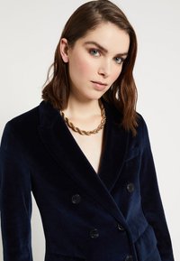 Tmavomodrý sametový dvouřadý blejzr s špičatými klopami a černými knoflíky. Hromadný zlatý řetězový náhrdelník doplňuje výstřih.