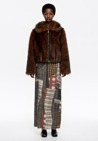 Chaqueta de borreguito marrón con cremallera y un cuello ancho. Combinada con una falda larga de patchwork que presenta varios patrones de cuadros en colores apagados.