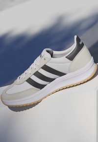 Weißer und grauer Adidas-Sneaker mit schwarzen Streifen, der auf einer weißen Oberfläche ruht und einen Schatten auf einen blau-weißen Hintergrund wirft.