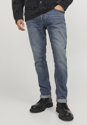Blugi din denim albastru închis, cu o croială slim, cusături vizibile și manșete răsucite, asortați cu cizme negre cu șireturi, pe un fundal neutru.
