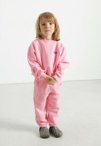 Ružová mikina a tepláky vyrobené z mäkkého materiálu, s malým modrým logom, skombinované so šedými slip-on teniskami. Hladká textúra a uvoľnený strih.