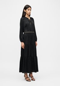 Μακρύ μαύρο φόρεμα με μακριά μανίκια, βολάν στο τελείωμα και κορδόνια στον λαιμό, με υφασμάτινο σχέδιο και μεταλλική ζώνη αλυσίδα. Μαύρες ψηλοτάκουνες σανδάλες με λουριά.