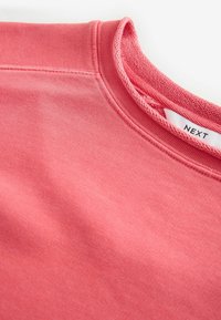 Koraalkleurige sweatshirt met een ronde hals, zachte textuur en gebreide accenten langs de kraag. Merkenlabel zichtbaar aan de binnenkant.