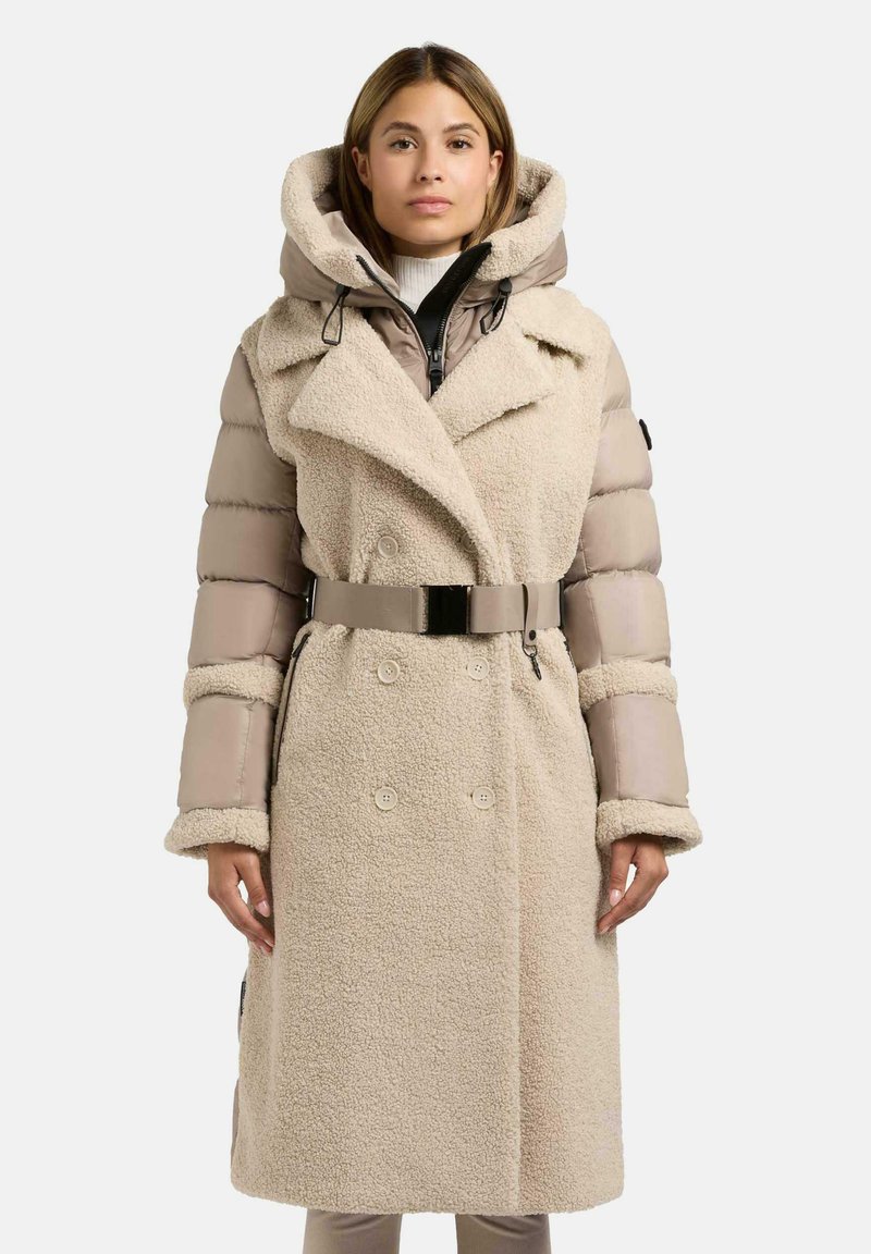 Beige dubbelzijdige jas met een gestructureerde schapenvacht en smooth, gewatteerde mouwen. Voorzien van een strakke riem en een grote capuchon met trekkoorden.