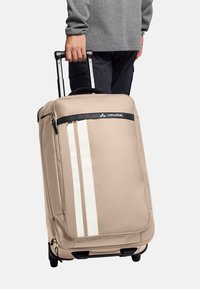 Beige koffer op wielen met een getextureerd oppervlak, voorzien van twee verticale witte strepen, een zwarte handgreep aan de bovenkant en een uitschuifbare zilveren handgreep.