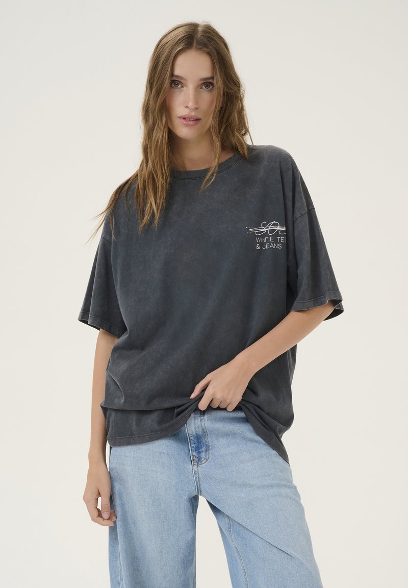 Tmavošedé oversized tričko vyrobené z mäkkého bavlneného materiálu, s jemne potlačeným logom na hrudi. Skombinované s svetlo modrými džínmi.