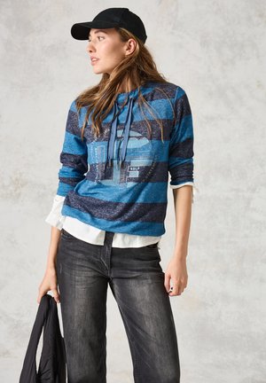 Blauwe en marine gestreepte sweatshirt met capuchon, voorzien van grafisch ontwerp. Combinatie met donkere jeans en een zwart jack in de hand, gedragen met een wit shirt.