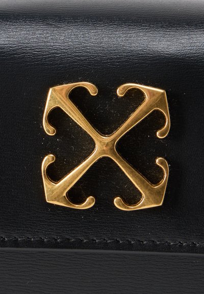 Λογότυπο από χρυσό μέταλλο σχηματισμένο από τέσσερα σταυρωμένα βέλη με καμπύλες άκρες σε μαύρο δερμάτινο φόντο με υφή.