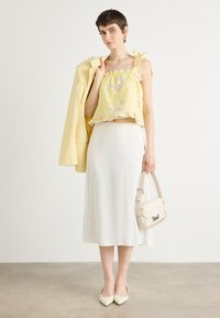 Top court jaune avec bord en dentelle, jupe midi blanche, blazer jaune clair et sac à main beige. Le modèle porte des talons blancs pointus.
