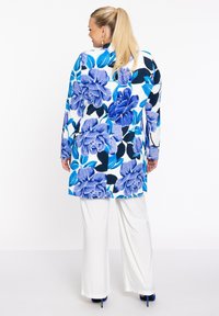 Blumenmuster langer Cardigan in Blau und Weiß, mit übergroßen Rosen und Blättern. Kombiniert mit weißen weit geschnittenen Hosen und blauen High Heels.