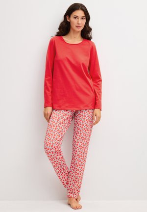 Vrouw met lang donker haar die een rood topje met lange mouwen en witte broek met een rood-roze fruitpatroon draagt, staat blootsvoets tegen een effen muur.