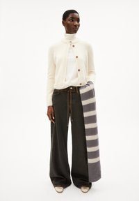 Cardigan color beige chiaro con scollatura rotonda e bottoni marroni, abbinato a jeans larghi scuri. Sciarpa grigia a righe poggiata su un braccio.