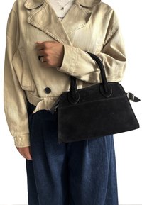 Černá semišová kabelka se strukturovaným tvarem, kulatými madly a dekorativním švem. Nositá s krémovým sáčkem a modrými kalhotami.