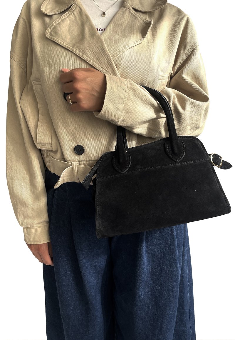 Černá semišová kabelka se strukturovaným tvarem, kulatými madly a dekorativním švem. Nositá s krémovým sáčkem a modrými kalhotami.