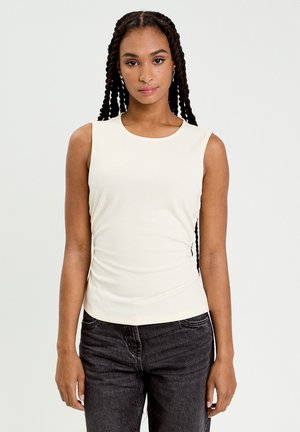 Giovane donna con lunghe trecce che indossa una canottiera bianca e jeans neri, in piedi davanti a uno sfondo chiaro e uniforme.