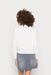 Witte langwerpige blouse met een relaxte pasvorm en subtiele textuur, gecombineerd met een grijze denimrok met een beschadigde zoom en twee achterzakken.