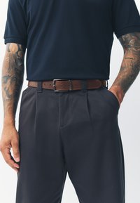 Bruine leren riem met zilverkleurige metalen gesp, gedragen bij een marineblauw poloshirt en antracietkleurige broek, met een gladde stof en een getailleerde pasvorm.