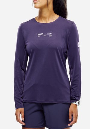 Frau trägt ein lila Langarm-Millet-Shirt mit weißen Logos auf Brust und Ärmel, dunkelblaue Shorts und eine weiße Armbanduhr.