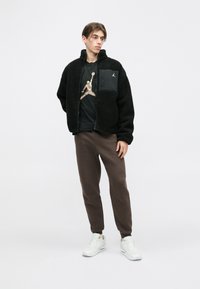 Fekete bolyhos dzseki cipzárral és oldalzsebbel, fekete grafikai póló, barna melegítőnadrág és fehér sportcipő. Egyszerű, lezser öltözet.