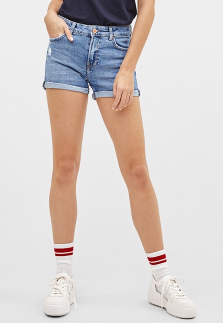 Pantalones cortos de mezclilla en azul claro, desgastados con dobladillos enrollados; combinados con zapatillas blancas y calcetines con rayas rojas. Diseño de cinturilla simple.