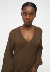 Maglione marrone a maglia con scollo a V profondo, maniche lunghe e un motivo testurizzato. Il tessuto sembra morbido e leggermente oversize.
