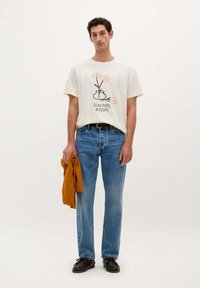 Crème kleurig shirt met korte mouwen en bloemenprint, voorzien van de tekst "SCALPERS BLOOM!", gecombineerd met lichtblauwe jeans en een mosterdkleurige trui vastgehouden. Zwarte schoenen.