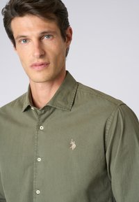 Camicia verde oliva con bottoni realizzata in cotone. Presenta un colletto, una patta frontale con bottoni e un piccolo logo ricamato sul petto.
