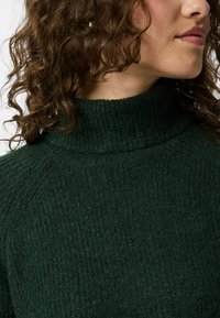Maglione lavorato a maglia verde scuro con un alto colletto a dolcevita e superficie testurizzata, caratterizzato da dettagli a coste lungo il collare.