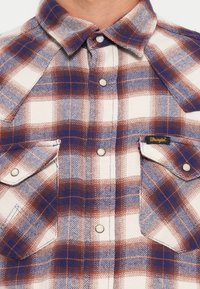 Camisa de cuadros de manga larga en tonos blanco, azul y rojo; presenta cuello tipo botón, dos bolsillos en el pecho y etiqueta de Wrangler en el costado.
