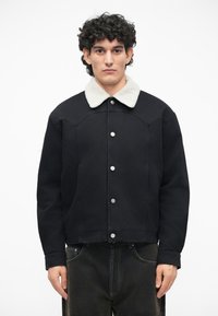Chaqueta de denim negra con cuello de pelaje sintético, que presenta botones a presión plateados y un diseño corto. Se lleva con jeans oscuros.
