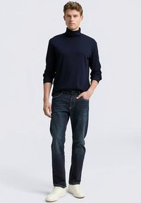 Marineblauer Rollkragenpullover, kombiniert mit dunklen Denim-Jeans. Das Model trägt weiße Sneaker. Der Pullover hat lange Ärmel, ein schlankes Design und eine figurbetonte Silhouette.