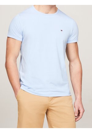Mand iført lyseblå T-shirt med korte ærmer og lille brystlogo, beige bukser, stående med den ene hånd i lommen mod neutral baggrund.