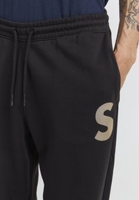 Zwarte trainingsbroek met een elastische tailleband en koord, met een groot, lichtgekleurde "S" logo op de linker bovenbeen.