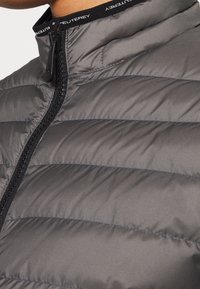 Grå pufferjakke med horisontal quiltet tekstur, udstyret med høj krave og sort lynlås. Mærkets logo synligt på indersiden af kraven.