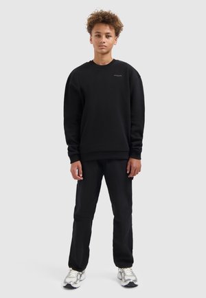 Zwarte sweatshirt met een ronde hals en geribde manchetten, gecombineerd met bijpassende zwarte broek. Model staat op zilveren en zwarte sneakers.