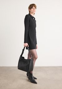 Bolsa de mão em couro preto com um design estruturado, textura suave e alça ajustável. A modelo veste um vestido preto de mangas longas e sapatos de salto agudo.